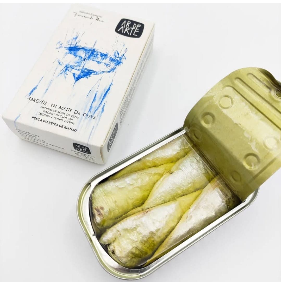 Open tin of sardines in olive oil beside its white and blue illustrated cardboard box.
