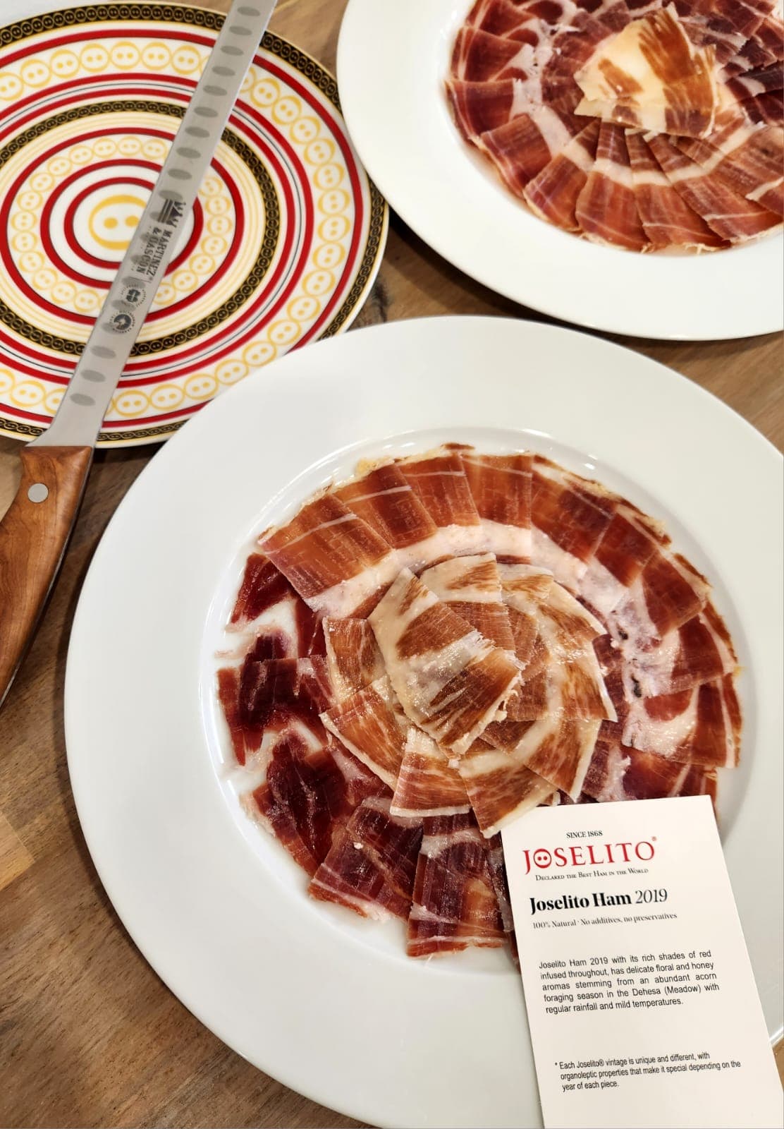 Thinly sliced Joselito ham on white plates with a long carving knife and product card.
