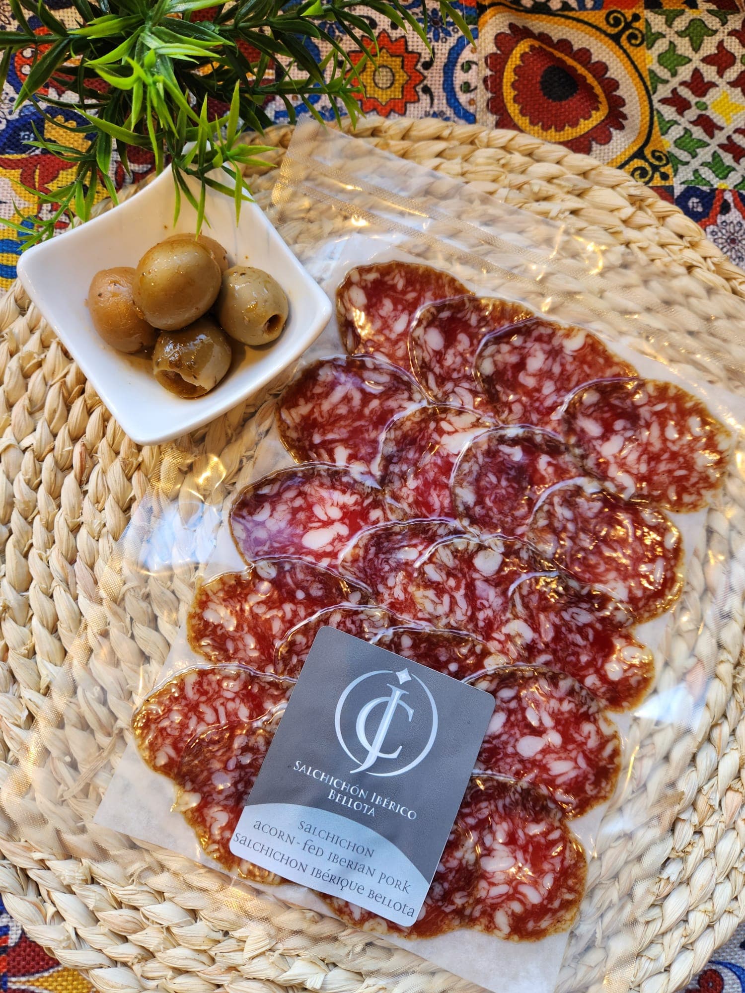 Sliced Salchichón Ibérico Bellota in vacuum packaging beside a small bowl of green olives.