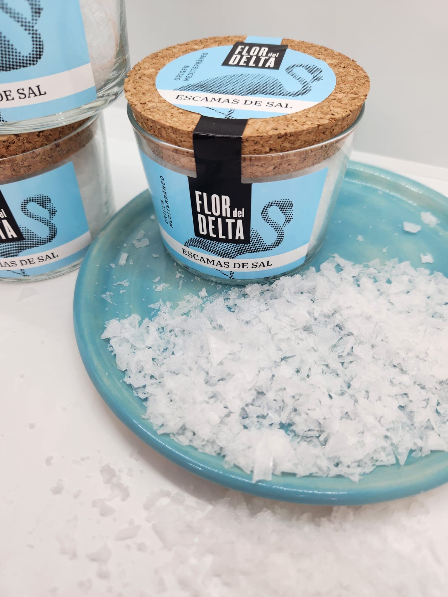 Jar of Flor del Delta sea salt flakes on a blue plate with scattered salt.